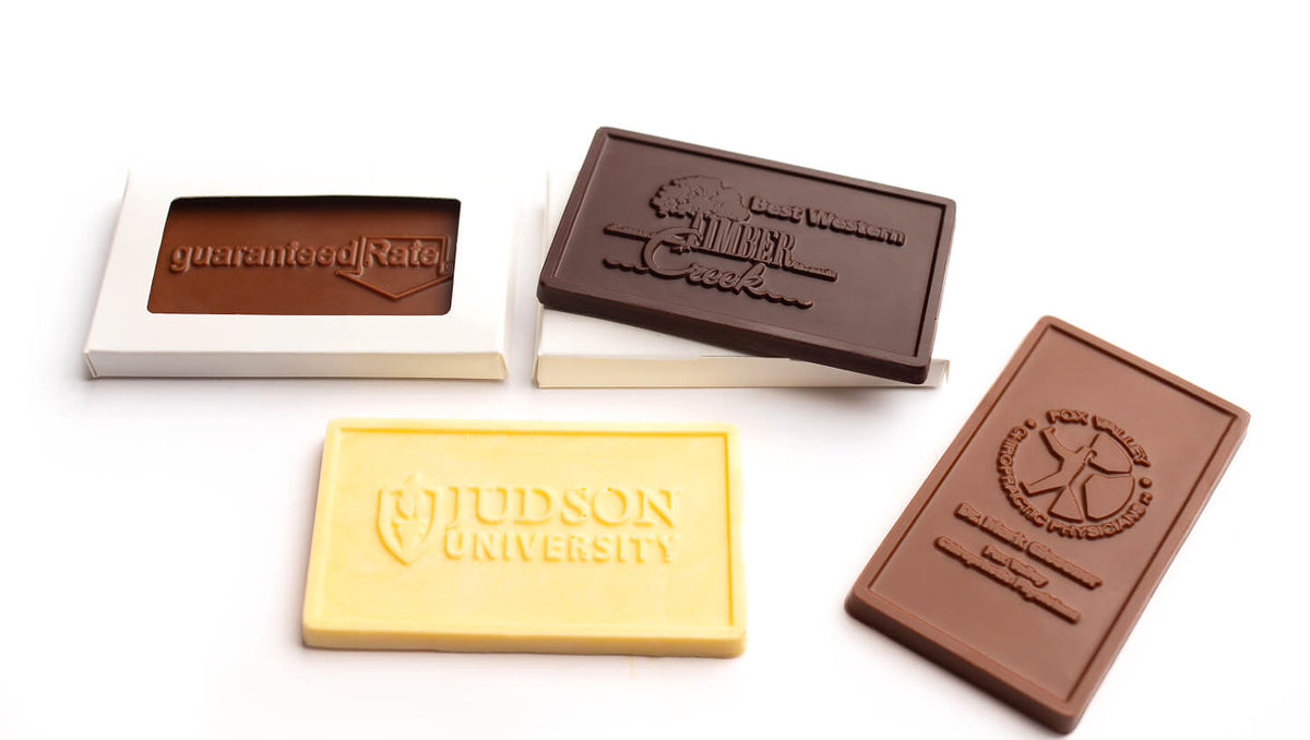 Four chocolate bars or chocolate-related pieces arranged on a white background: a dark brown chocolate bar in a white wrapper, a dark brown chocolate bar with an embossed logo, a pale yellow chocolate bar embossed with 'JUDSON UNIVERSITY,' and a medium brown chocolate bar with an embossed circular logo.