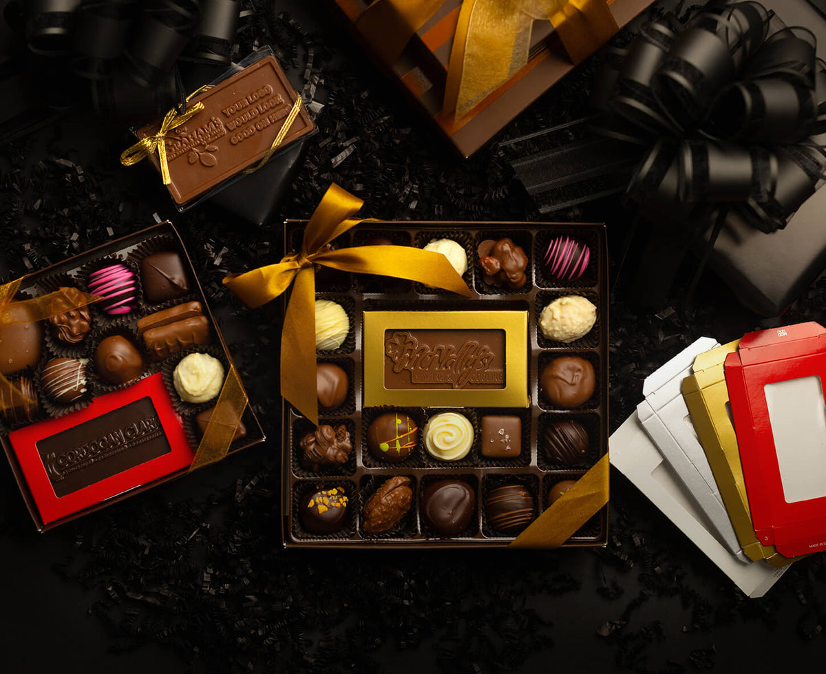 An assortment of gourmet chocolates in open gift boxes with gold and red packaging, arranged on a dark textured surface with decorative black ribbons and gift boxes around them.