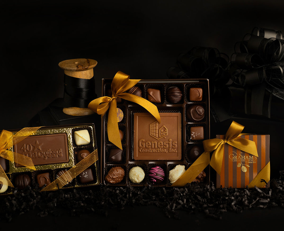 A luxury chocolate gift set featuring multiple boxes of assorted chocolates and truffles from Genesis Construction Co., styled with bright yellow ribbons on a dark background with shredded cocoa beans.