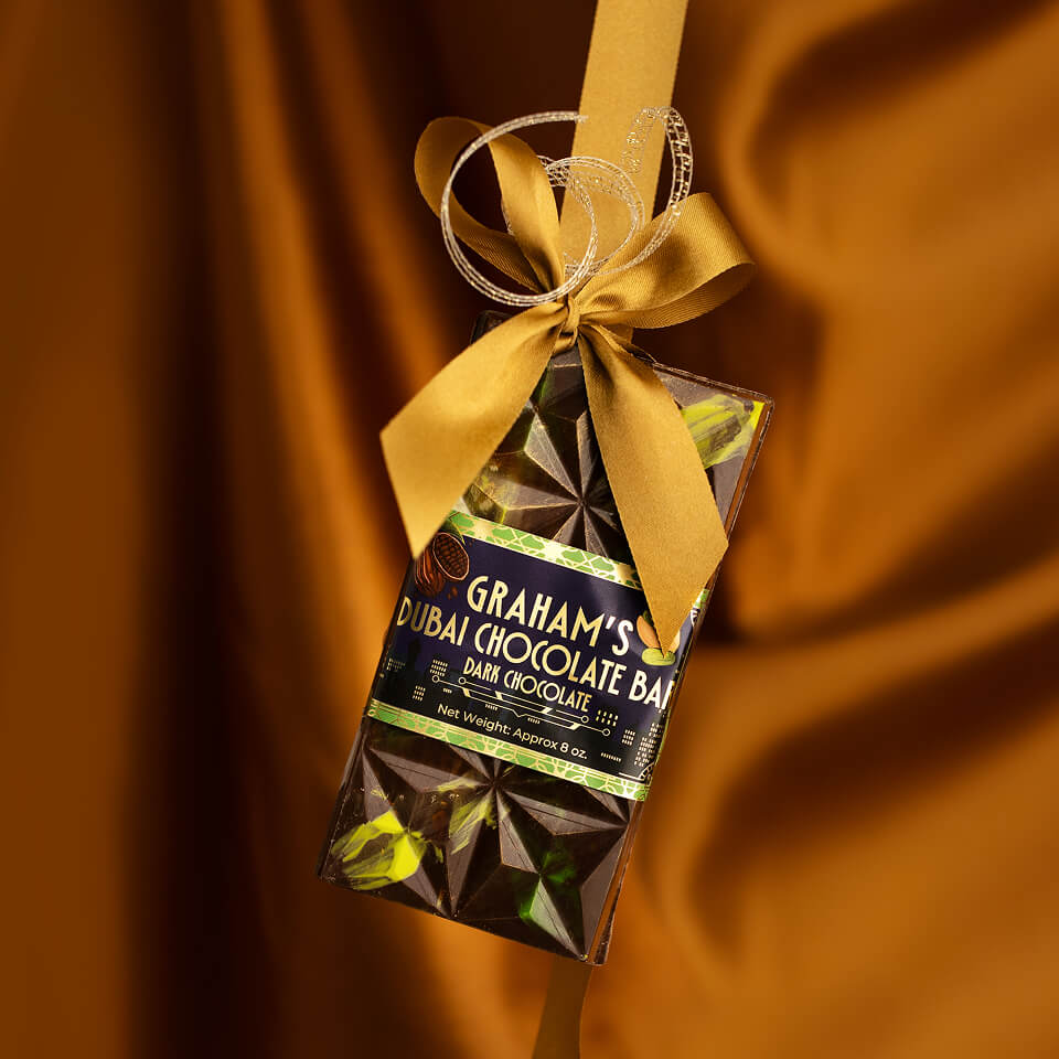 A packaged chocolate bar hanging from a gold ribbon bow against a draped warm brown background. The wrapper shows the brand name Graham's and reads 'Dubai Chocolate Bar Dark Chocolate'.