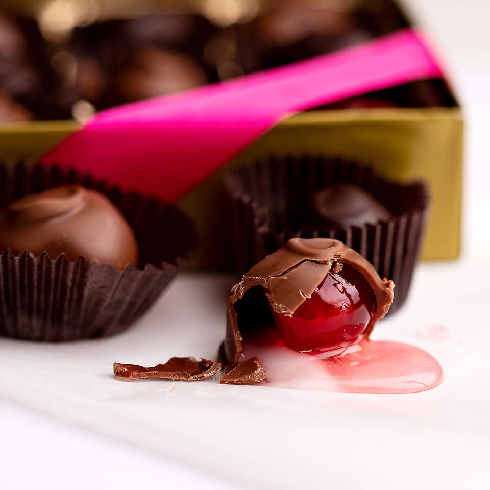 Chocolate truffles in dark brown paper cups, with one broken in front to reveal a glossy red cherry filling and a pink sugar glaze, set in front of an open box of chocolates with a bright pink ribbon.