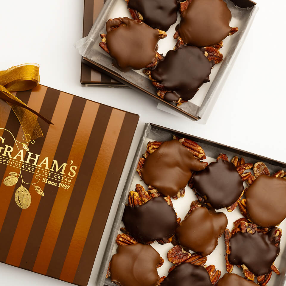 Two open boxes of Graham's Chocolates and Rice Cream, with one box closed and decorated with a gold ribbon. The chocolates are topped with dark chocolate and pecans, set against a clean white background.