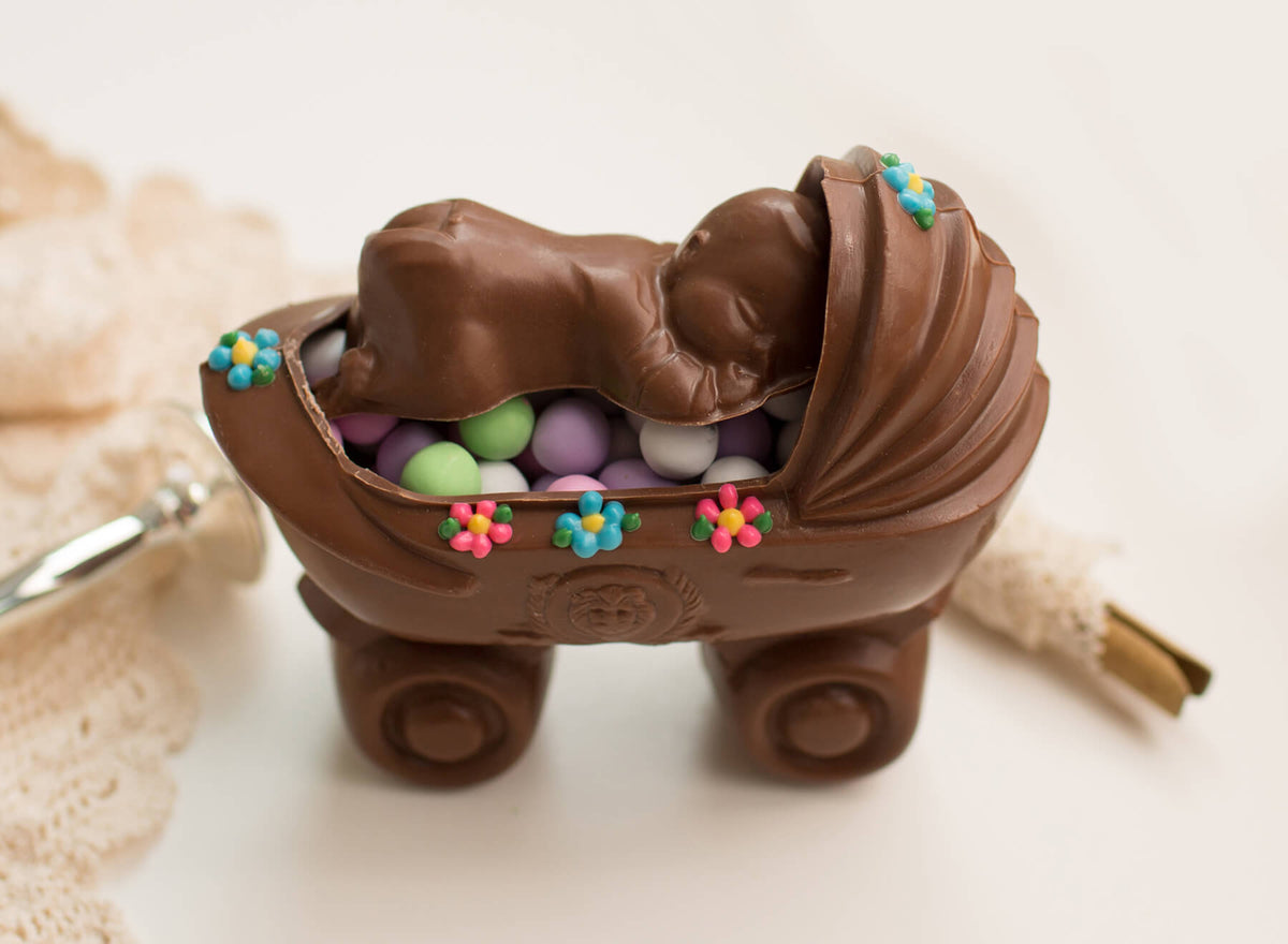 A chocolate baby carriage keychain with a sleeping baby figure inside, filled with colorful candy-coated chocolates and decorated with small flower accents, shown on a light background.