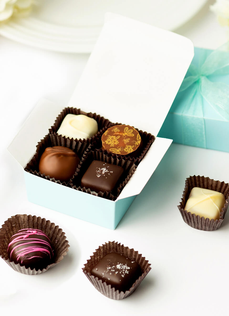 An open light blue box of assorted chocolates on a white surface, with three additional chocolates placed around it and a closed matching gift box in the background.