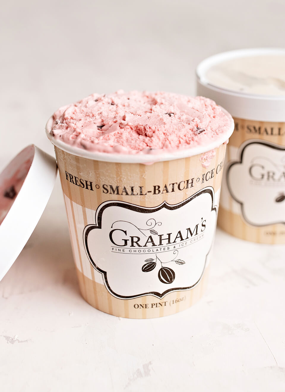 A pint of Graham's Fine Chocolate Ice Cream with pink strawberry swirl visible at the top, shown in a kraft paper cup with the lid partly off, on a light neutral background with another pint blurred in the back.