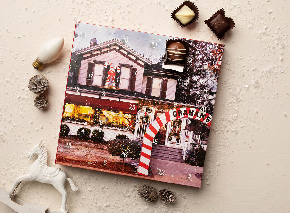 A Christmas advent calendar with a printed illustration of a house and numbered doors, styled on a light textured surface with chocolates, pinecones, a white horse figurine, and a light bulb.
