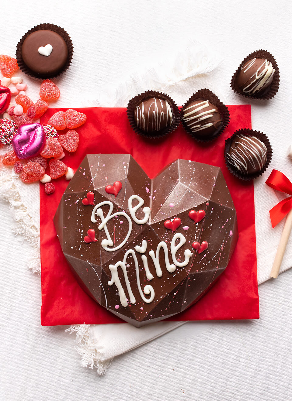 A heart-shaped chocolate gift box with the words 'Be Mine' in white icing, surrounded by assorted chocolates, candy, and red wrapping paper on a white surface.