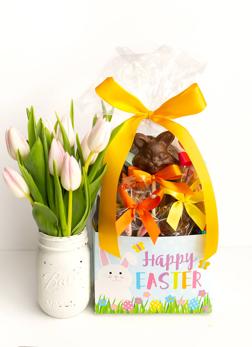 An Easter gift set featuring a box of chocolate treats with a bunny face on the front, wrapped in clear cellophane with a large yellow ribbon, next to a white mason jar filled with pink tulips on a white background.