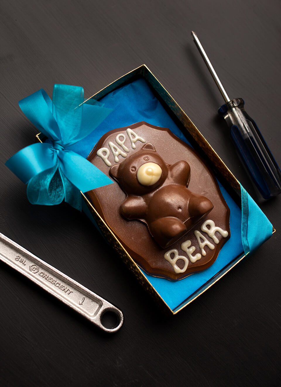 A chocolate bear-shaped gift box with the words 'PAPA BEAR' in white icing, presented in an open box with a bright blue ribbon. The box is styled on a dark background with wrench and screwdriver props.