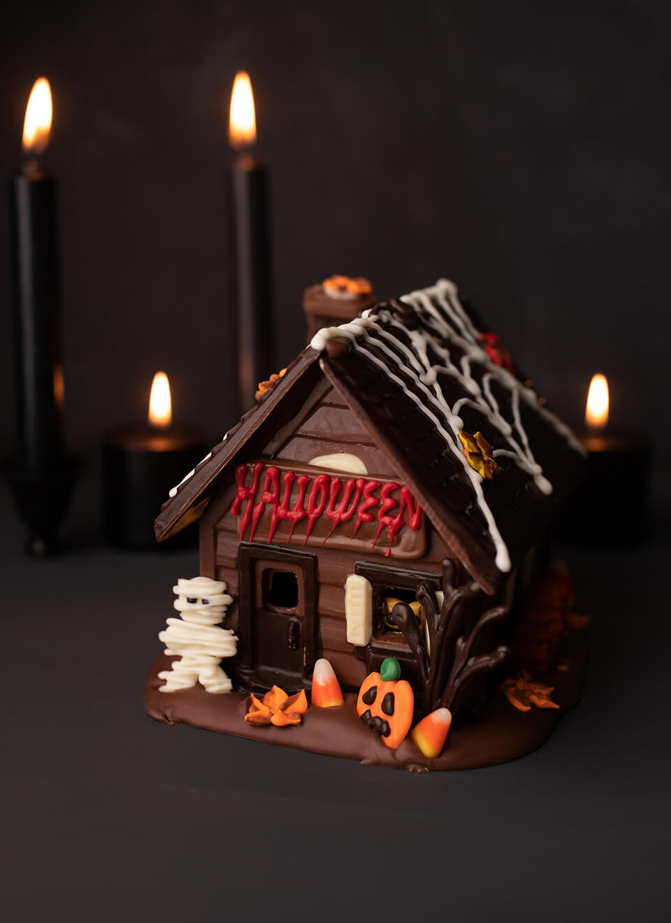A Halloween-themed miniature house decoration or cake, decorated with dark brown icing, white spiderwebs, red 'HALLOWEEN' lettering, a white mummy figure, candy corn, and orange leaves, set against a dark background with lit black candles.