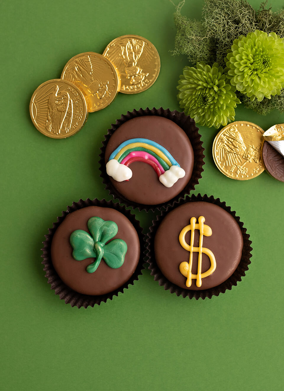 Three chocolate cupcakes decorated with St. Patrick's Day themed icing designs, arranged on a green background with gold foil coins and green flowers.