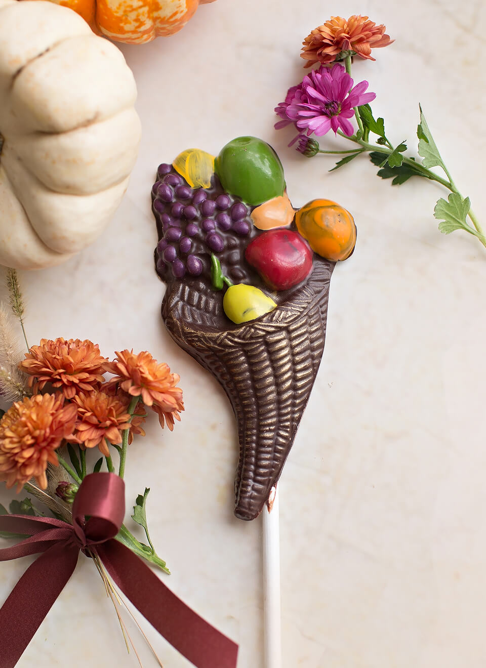A decorative chocolate cornucopia lollipop with colorful fruit details, styled on a light marble surface with autumn flowers, a white pumpkin, and a dark ribbon.
