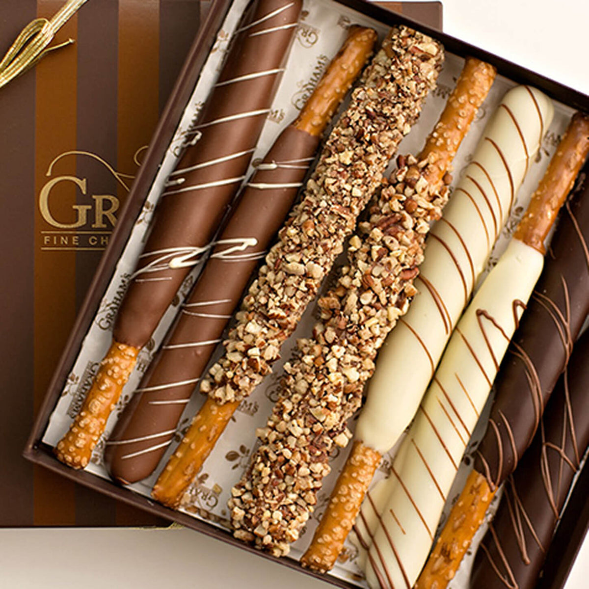 An open box of assorted chocolate-covered pretzel rods, including milk chocolate, dark chocolate, white chocolate, and nut-coated varieties, with a branded brown gift box visible beside them.