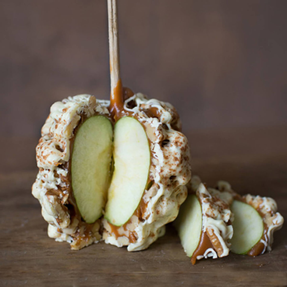 A close-up of a caramel apple skewer with two green apple slices, coated in white chocolate and drizzled with caramel sauce, on a wooden surface.
