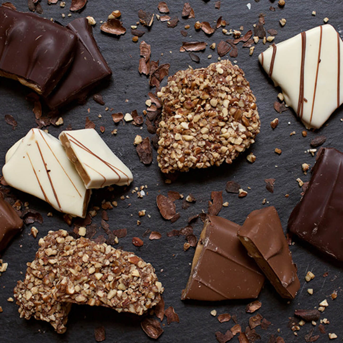 An assortment of gourmet chocolate pieces and nut-coated treats arranged on a dark slate background, including dark chocolate pieces, milk chocolate chunks, white chocolate squares with drizzle, and two rough nut-coated chocolate pieces.