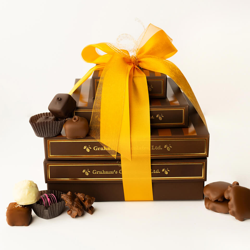 A stack of gourmet chocolate boxes tied with a large yellow ribbon, with assorted chocolates placed around and on top of the boxes against a clean white background. The boxes are dark brown with gold lettering that reads 'Gruau's Chocolate Co.