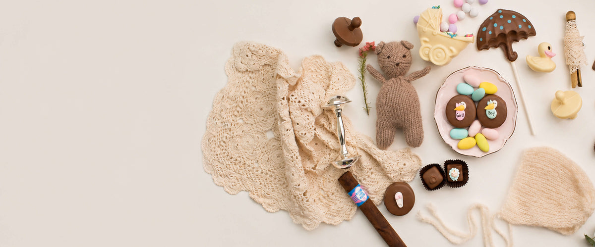 A styled flat-lay of a crocheted baby blanket and matching accessories, including a teddy bear, small crocheted toys, candy decorations, and a pacifier clip, arranged on a light beige background.