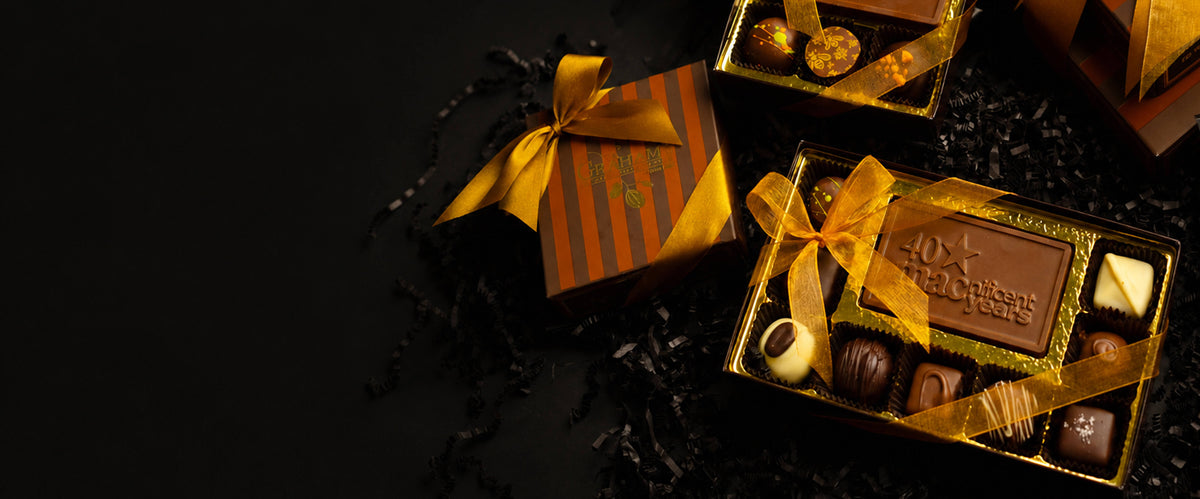 An open box of assorted chocolates tied with a gold ribbon sits on a black background beside a striped gold and brown gift box, with more chocolate boxes partially visible at the top right.