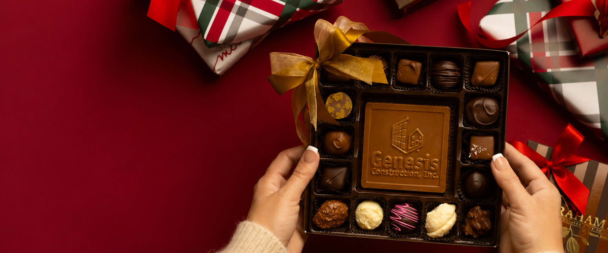 A top-down view of a person holding a box of assorted chocolates, including a central square milk chocolate bar embossed with the Genesis Chocolate Company logo. The box is decorated with a gold ribbon and set against a dark red background with wrapped gifts in the corners.
