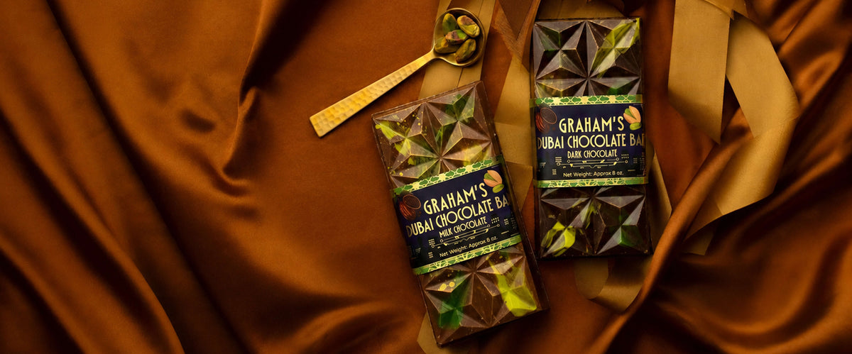 Two bars of Graham's Urban Chocolate Bar in dark chocolate flavor, displayed on a draped brown satin fabric with a small gold spoon.