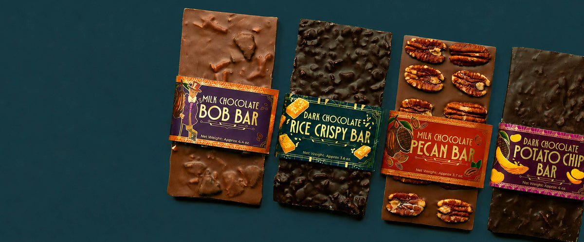 Four chocolate bars arranged upright on a dark teal background, each with a colorful paper wrapper. The wrappers read Milk Chocolate Bob Bar, Dark Chocolate Rice Crispy Bar, Milk Chocolate Pecan Bar, and Milk Chocolate Potato Chip Bar.