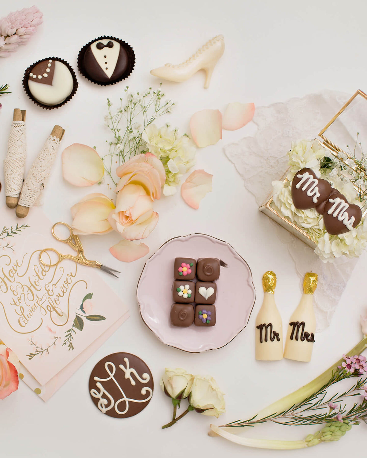 A styled flat-lay of wedding-themed chocolate treats and decorations on a white surface, including chocolate cupcakes shaped like a bride and groom, small chocolate squares with heart and flower centers, two cream champagne bottle-shaped chocolates with gold tops and dark lettering, a small box of white flowers with heart-shaped chocolates reading 'Mr. & Mrs.