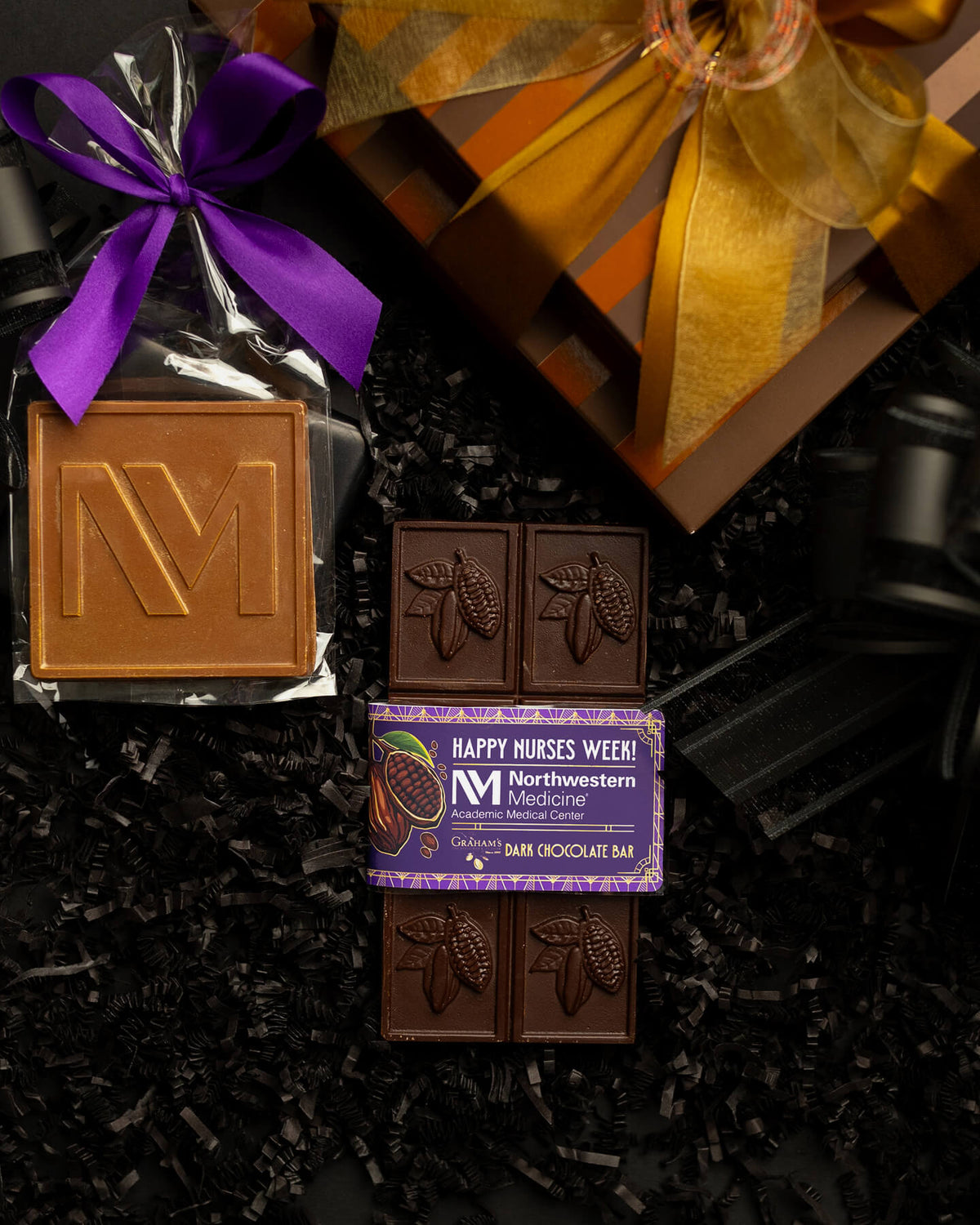 A chocolate gift set arranged on black shredded paper, featuring a wrapped milk chocolate bar with a purple ribbon, a boxed dark chocolate bar labeled 'Masternose Medicine' with cocoa pod artwork, and a wrapped square milk chocolate bar with an embossed letter M. A gold ribbon and gift box are partially visible in the background.