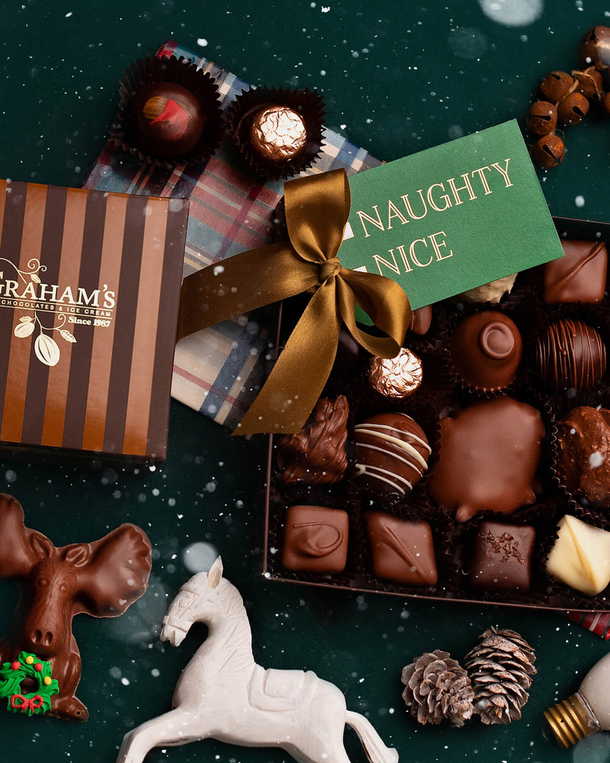 A festive assortment of chocolates and truffles arranged on a dark green snowy surface, with a striped Graham's box, a green card reading 'NAUGHTY NICE,' a gold ribbon, and holiday props including pinecones, a white horse figurine, and a light bulb.