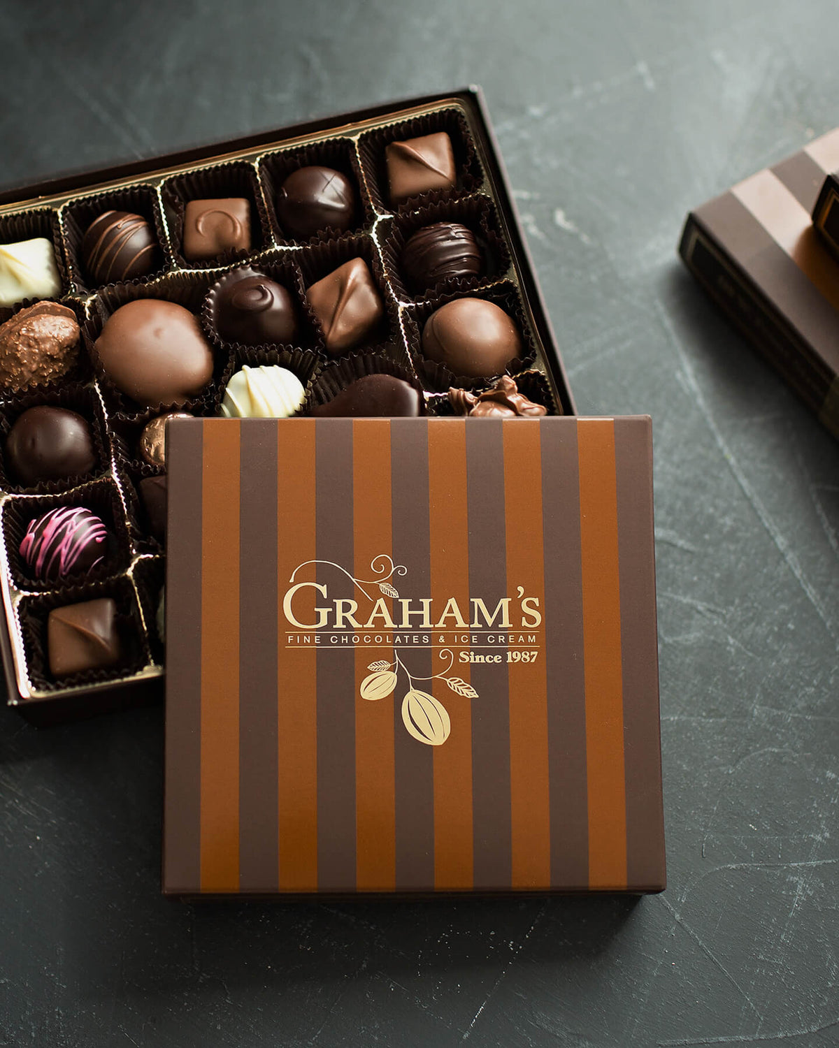 A premium chocolate gift box with brown and dark brown vertical stripes, featuring the brand name 'Graham's Fine Chocolates & Candy' in gold. The lid is placed in front of an open box displaying an assortment of milk, dark, and white chocolates on a dark slate surface.