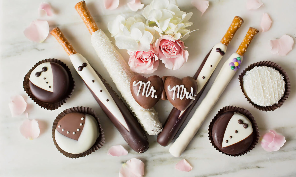 Bride and groom themed chocolate treats on a light marble surface, including decorated chocolate-covered pretzels, cupcakes, and heart-shaped chocolates spelling 'Mr. & Mrs.