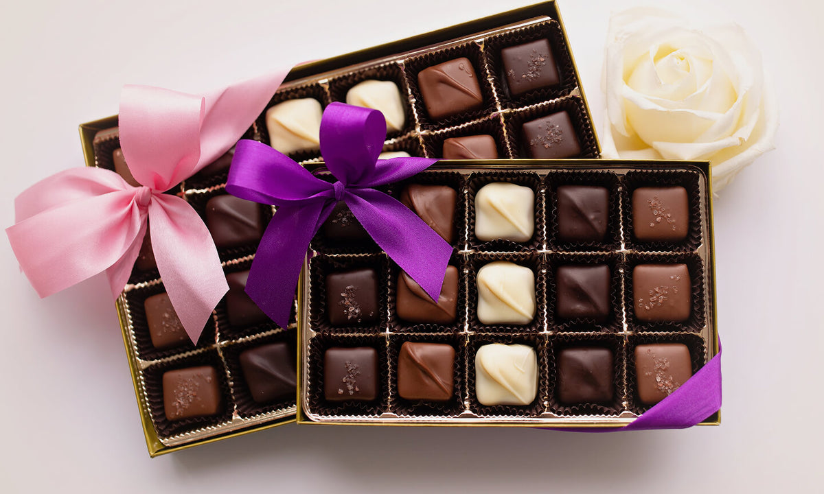 Two open boxes of assorted chocolates on a white background, one tied with a pink ribbon and the other with a purple ribbon, with a white rose in the upper right corner.