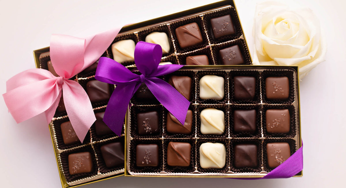Two boxes of assorted chocolates tied with pink and purple ribbons, arranged on a white background with a white rose prop.