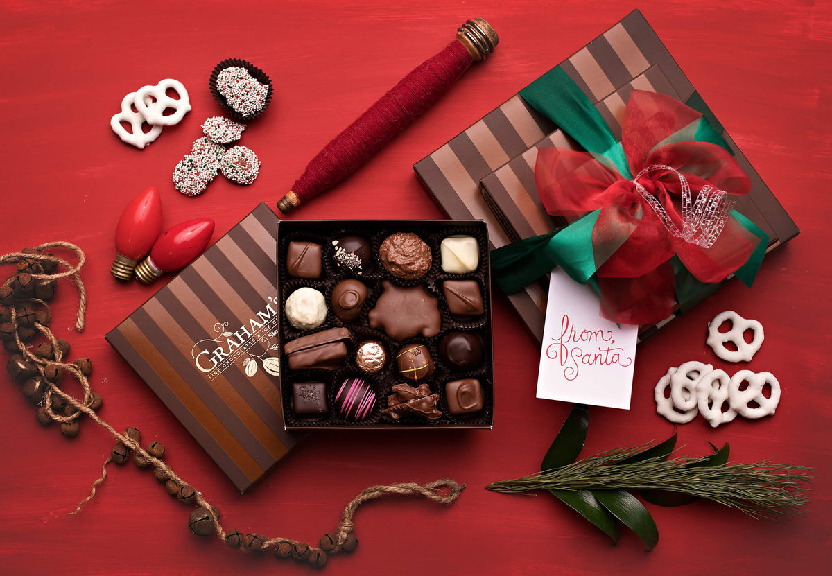 A gourmet chocolate gift box set displayed on a red background, featuring an open box of assorted chocolates, a wrapped gift box with a red bow and green ribbon, pretzels, red Christmas bulbs, pinecones, and a red candlestick.