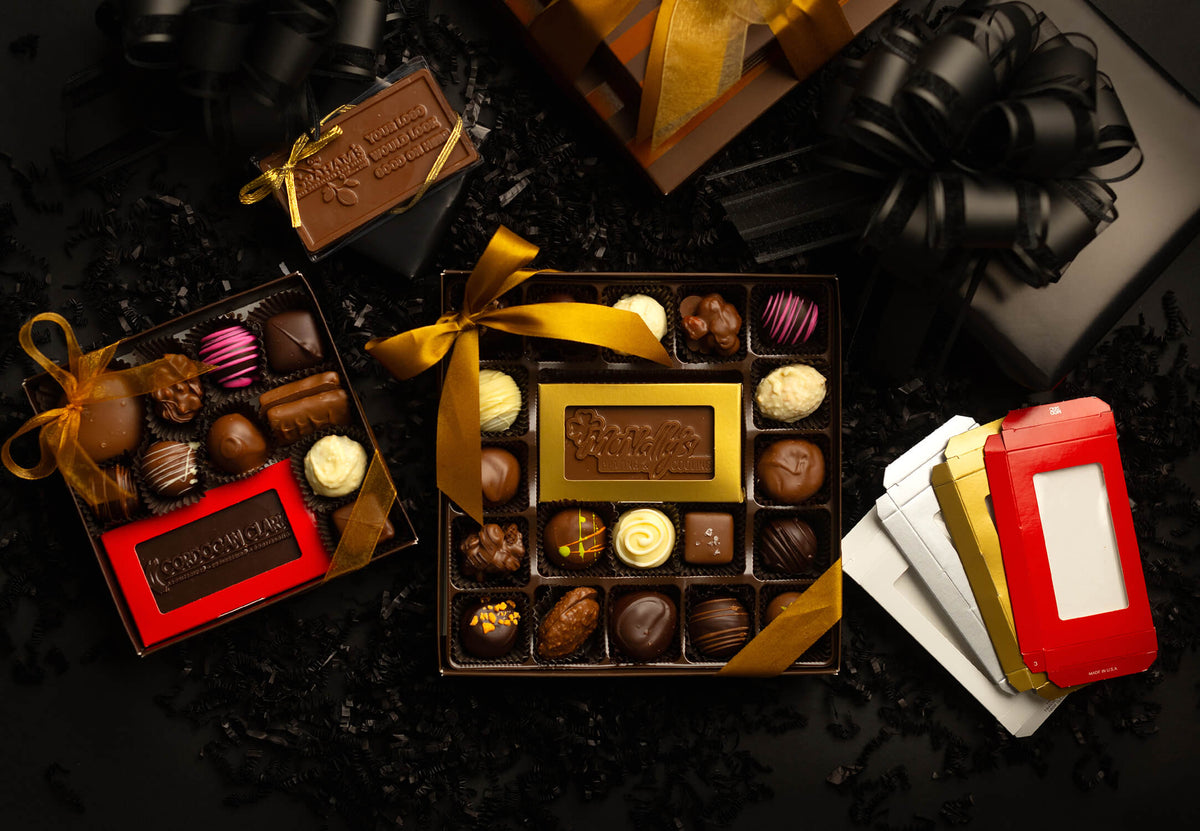 An overhead flat-lay of assorted chocolates in open gift boxes with gold and red packaging, surrounded by black ribbon bows and dark decorative filler on a black background.