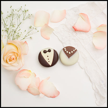 Pearl Necklace Bride + Tuxedo Groom. Two decorated chocolate Oreos shaped like a groom's tuxedo and a bride's wedding dress, styled on a white surface with pink rose petals, a peach rose, baby's breath, and lace.