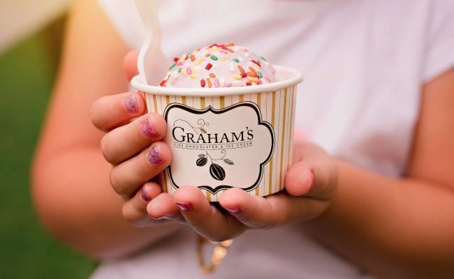 A close-up of a person holding a paper cup of Graham's gourmet ice cream or gelato outdoors. The cup is white with gold stripes and a prominent black and white label reading 'GRAHAM'S'.