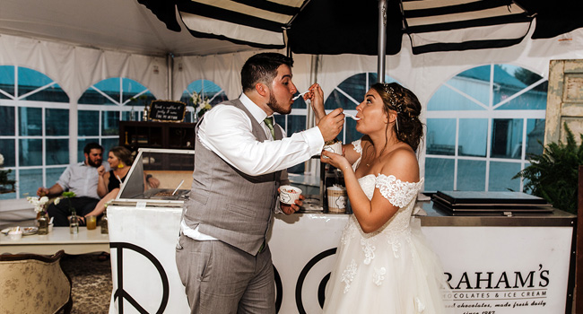 A bride in a white lace off-the-shoulder wedding gown and a groom in a gray vest and white shirt feed each other ice cream at a wedding reception under a black and white striped tent canopy. Graham's Chocolate Shop branding is visible on the counter.