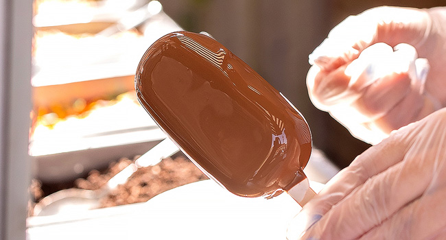 A glossy brown chocolate-covered ice cream bar on a stick, held by hands wearing clear disposable gloves in a brightly lit setting.