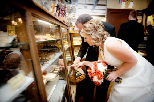 A bride in a white wedding gown and a man in a dark suit lean over a bakery display case, pointing inside. The bride holds a bouquet of red and white roses.