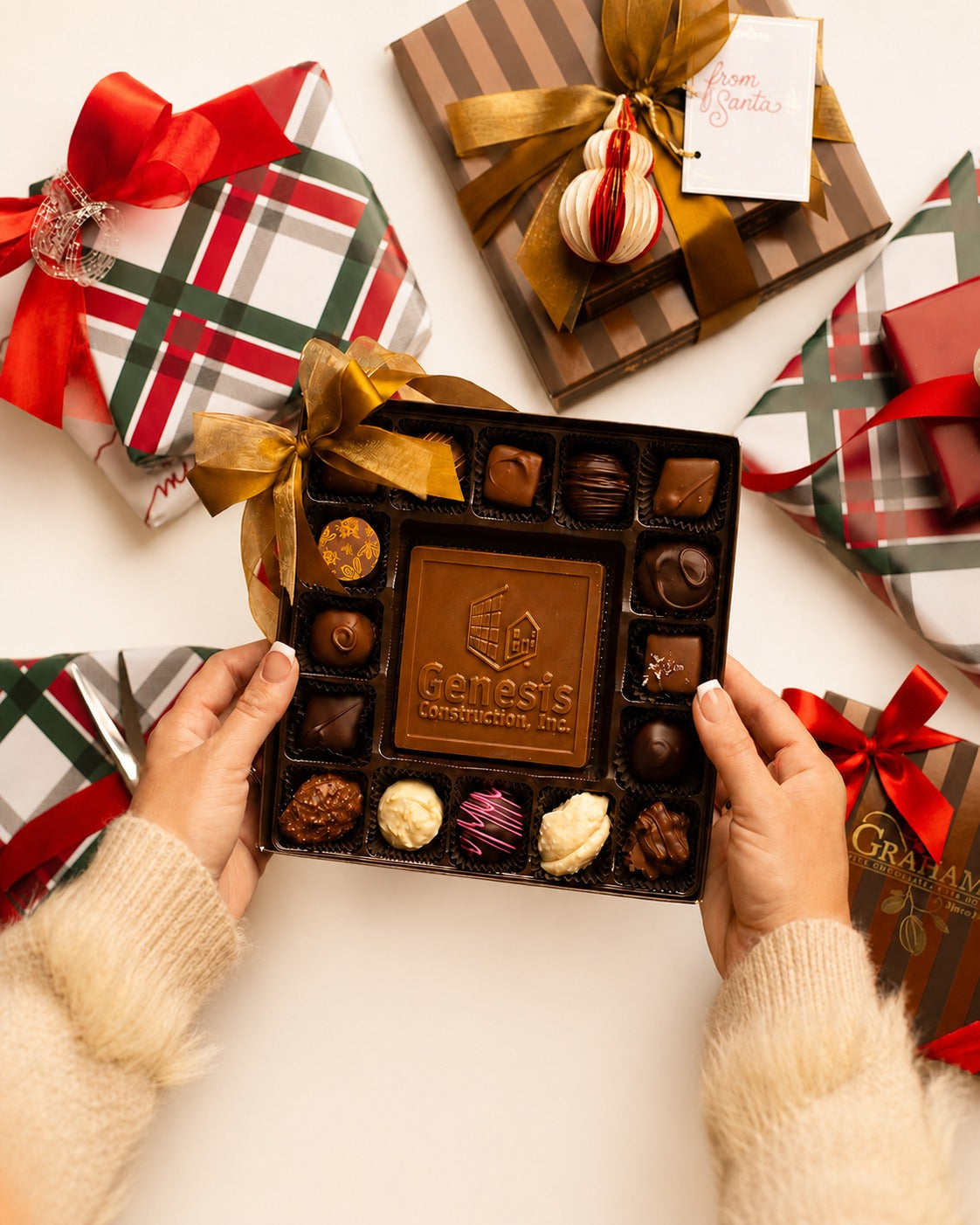 A top-down view of hands holding an open box of assorted chocolates, surrounded by wrapped holiday gift boxes. The center box is dark brown and features a gold logo that reads 'Genesis Confections Inc.