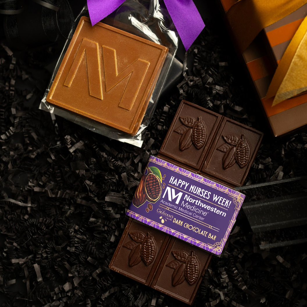 A gift assortment of chocolate bars and a wrapped square chocolate bar arranged on black shredded paper. The set includes a purple-wrapped chocolate bar with a large embossed letter M, a dark chocolate bar with cocoa pod embossing, a wrapped chocolate bar with a purple label reading 'Happy Nurses Week! Northwestern Medicine Center,' and part of an orange-and-brown gift box visible at the right edge.