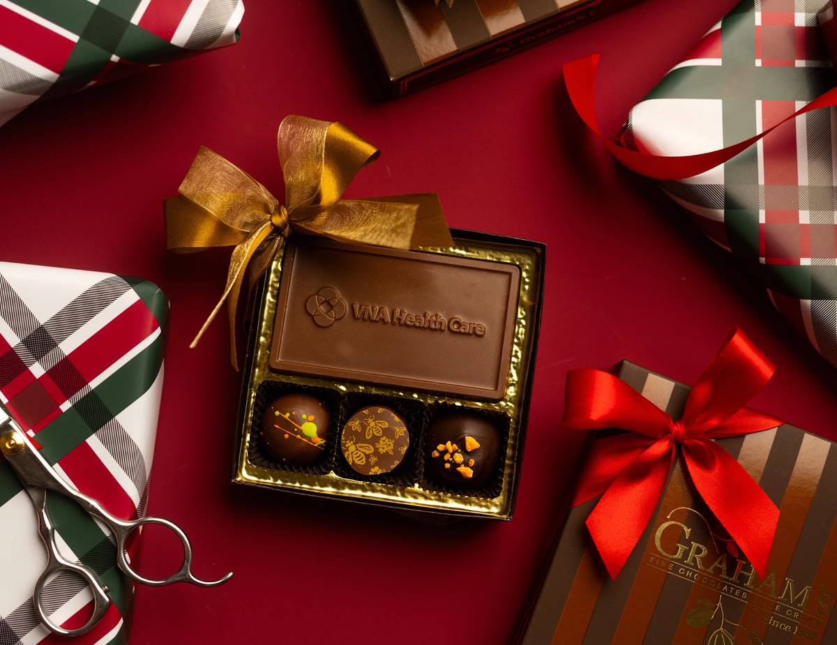 An open square chocolate gift box containing three decorated chocolates, tied with a gold ribbon, arranged on a dark red surface with wrapped presents and ribbon scissors around it. The chocolate bar reads 'L.