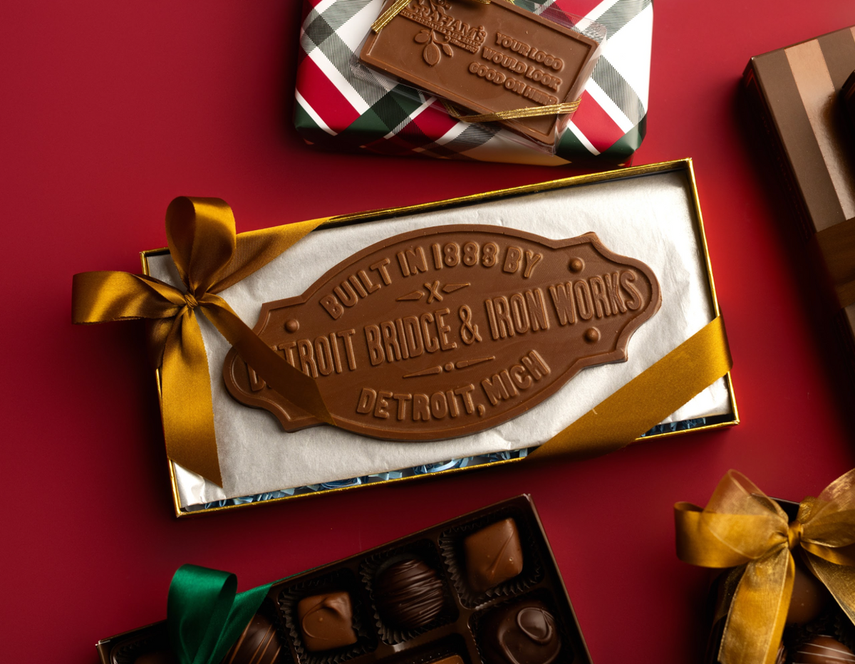 A boxed chocolate gift set on a red background, with a large chocolate plaque reading 'Pittsberry Detroit Bridge & Rail Works Detroit, Michigan' wrapped in a gold ribbon. Other chocolate boxes and wrapped gifts are partially visible around it.