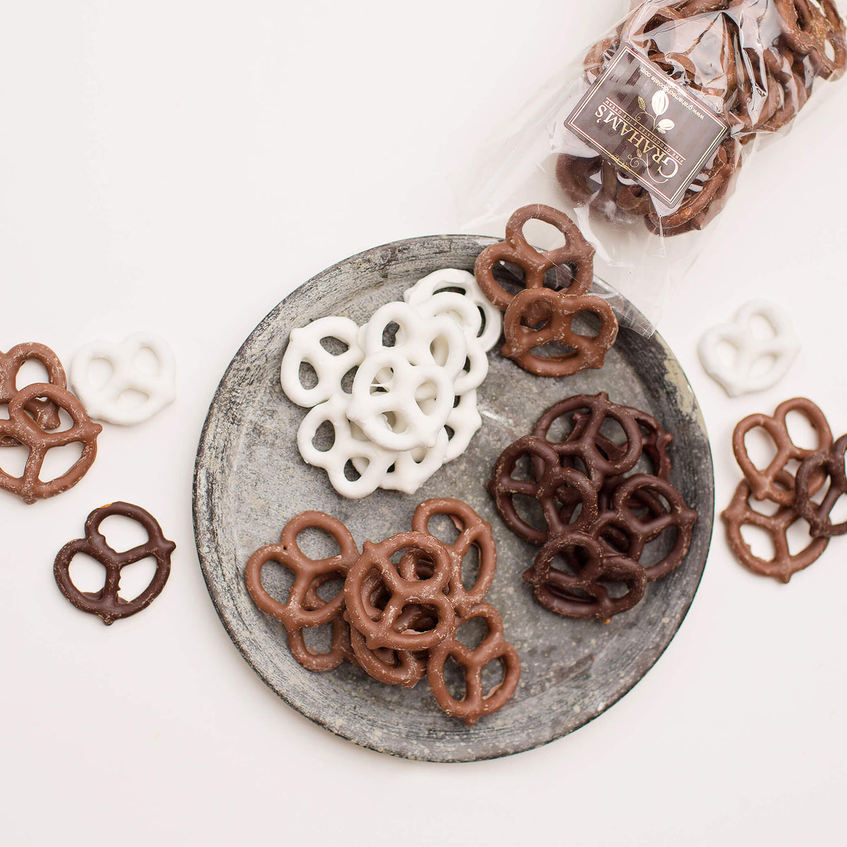 Chocolate pretzel snacks arranged on a round gray plate, with a small clear bag of pretzels in the upper right on a white background.