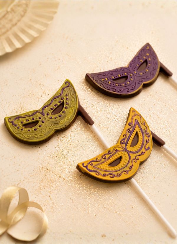 Three decorative masquerade mask lollipops on white sticks, featuring purple and gold, olive green and gold, and purple and brown designs, arranged on a glittery beige surface.
