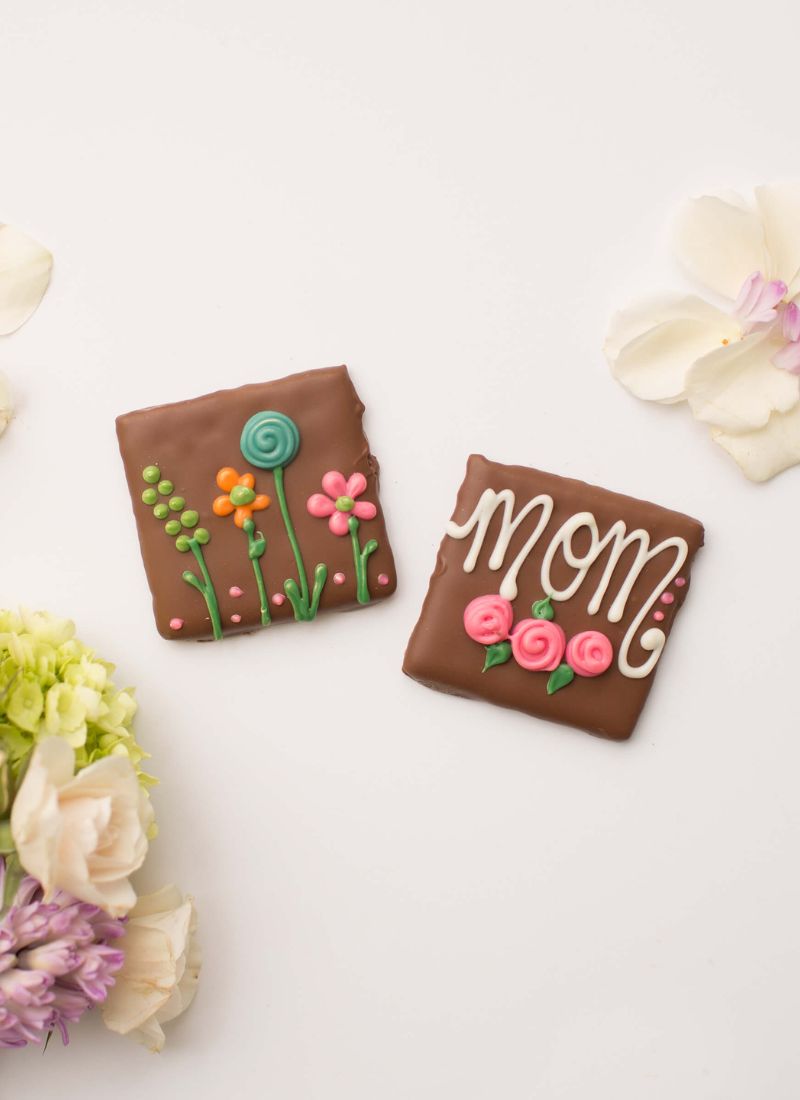 Two decorated square chocolate cookies on a white background with floral props. One cookie has colorful iced flowers and a lollipop design, and the other says 'mom' in white script with pink roses.