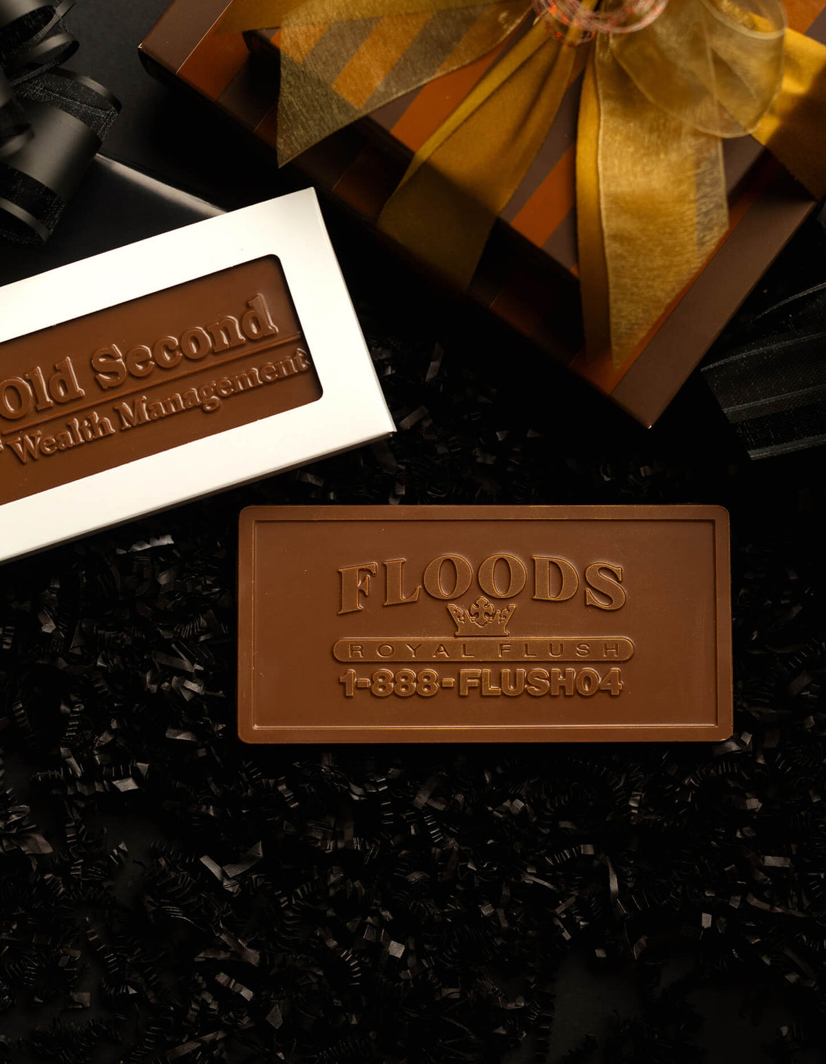 Two branded chocolate bars displayed on a black textured surface with shredded black filler. One bar is in a white box labeled 'Old Second Wealth Management,' and the other is a larger plain brown bar embossed with 'FLOODS REAL ESTATE' and a phone number.