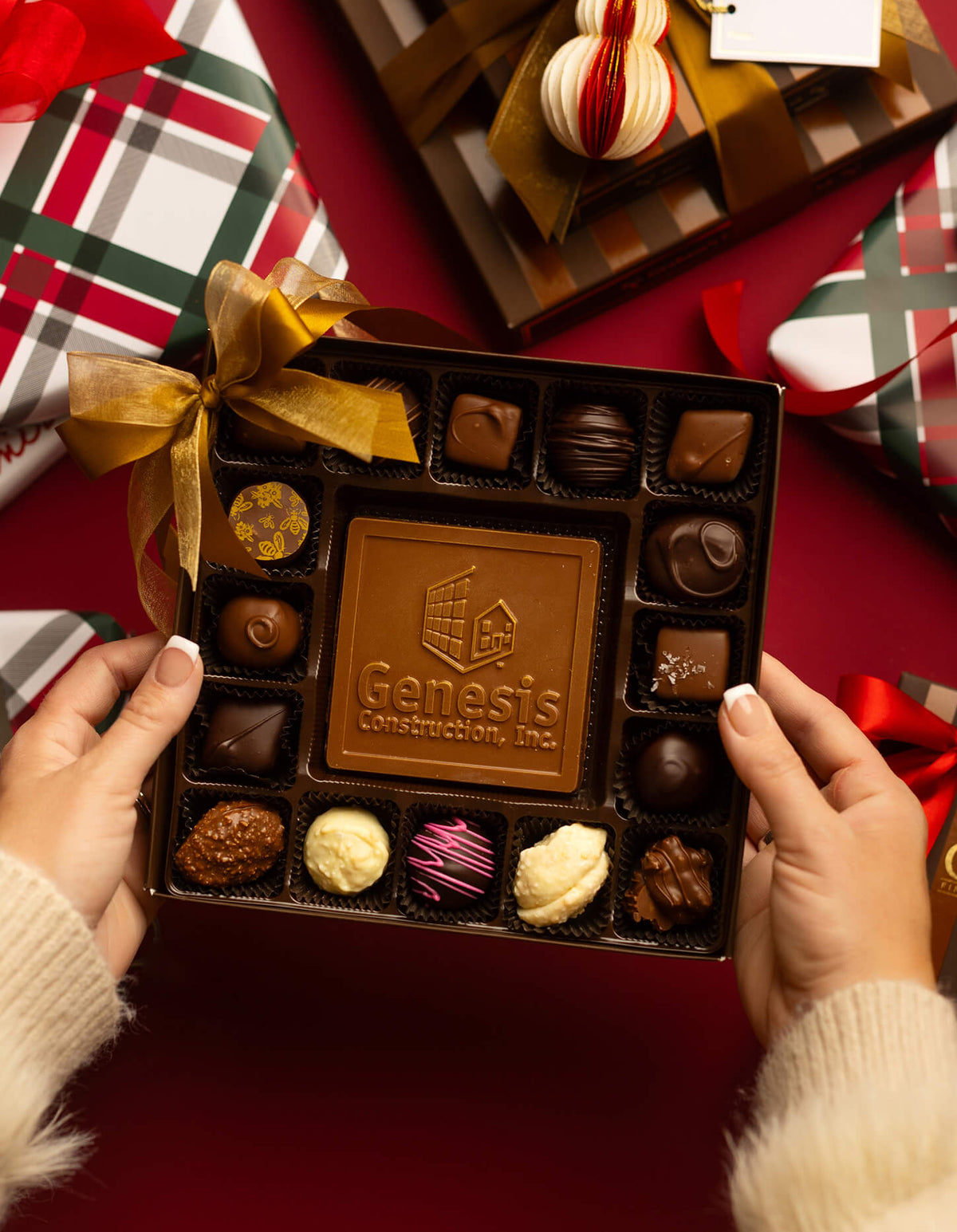 A person holds an open box of assorted chocolates, including a large center milk chocolate bar embossed with the Genesis Construction, Inc. logo.