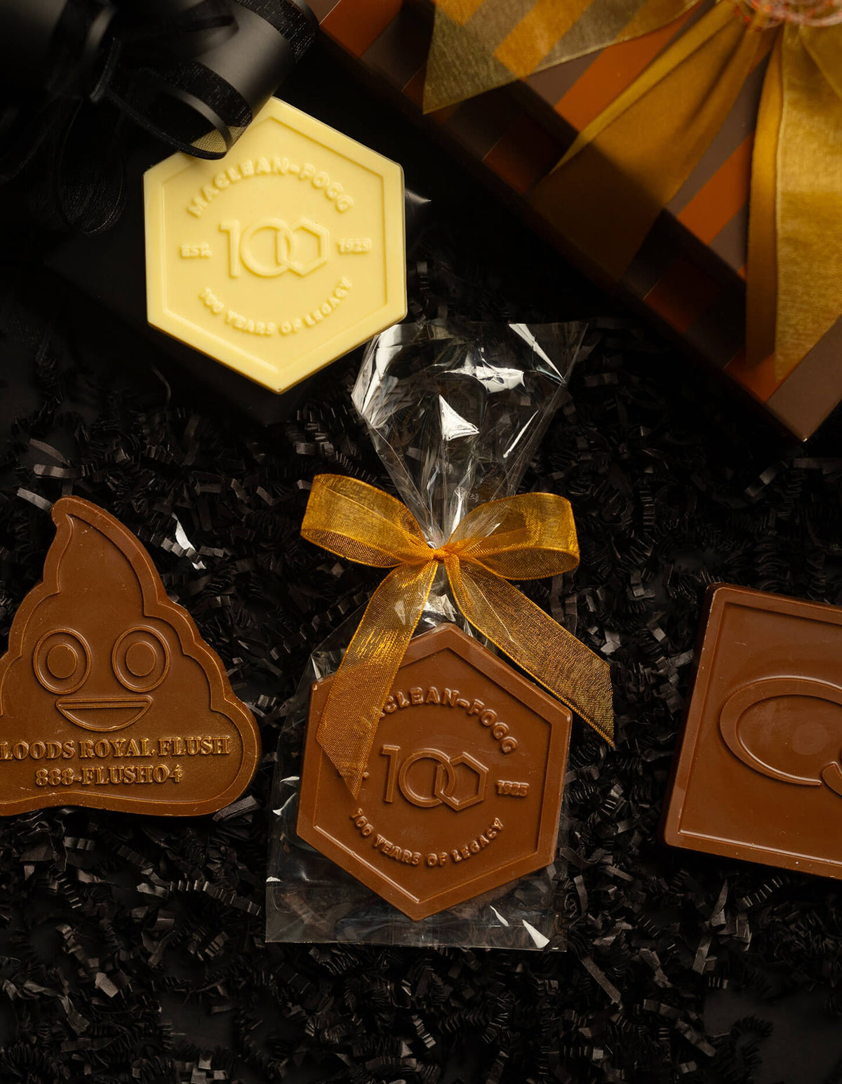 A gift assortment of custom chocolate treats from Candy Cane Company, featuring a pale yellow hexagonal chocolate, a brown hexagonal chocolate in clear wrapping with a gold ribbon, a brown poop-shaped chocolate, and a brown square chocolate, arranged on black shredded paper with a gift box in the background.