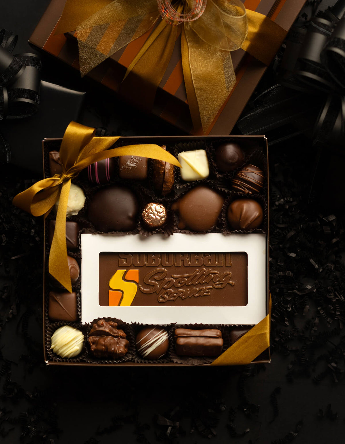 An open box of assorted chocolates with a branded chocolate bar in the center, tied with a gold ribbon, photographed on a dark background with a matching gift box above.